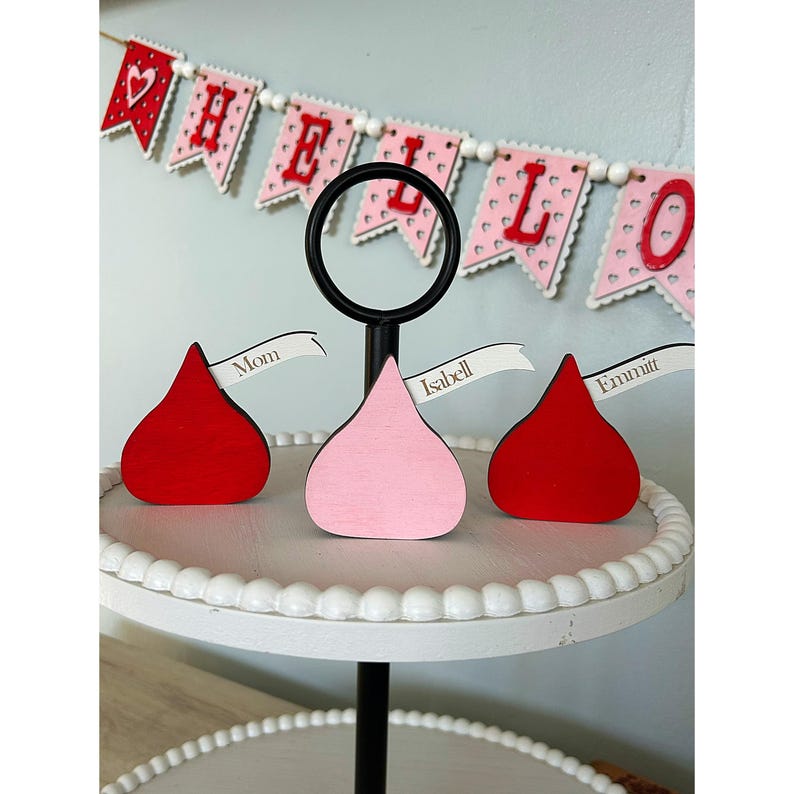 May include: Three wooden chocolate kiss-shaped ornaments with personalized names on white ribbon banners. The ornaments are red, pink, and red and are displayed on a white tiered cake stand with a black base. The background features a red and white "Hello" banner.