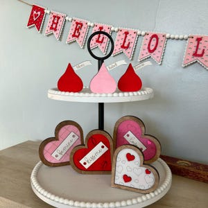May include: A two-tiered white tiered tray with a black metal stand. The top tier has three red, pink, and white wooden heart-shaped ornaments with the words "Mom", "Isabel", and "Emma" written on them. The bottom tier has four wooden heart-shaped ornaments with the words "Love", "Kiss Me", "Bemine", and a heart with a pattern of small hearts. The tray is decorated with white pearl beads.