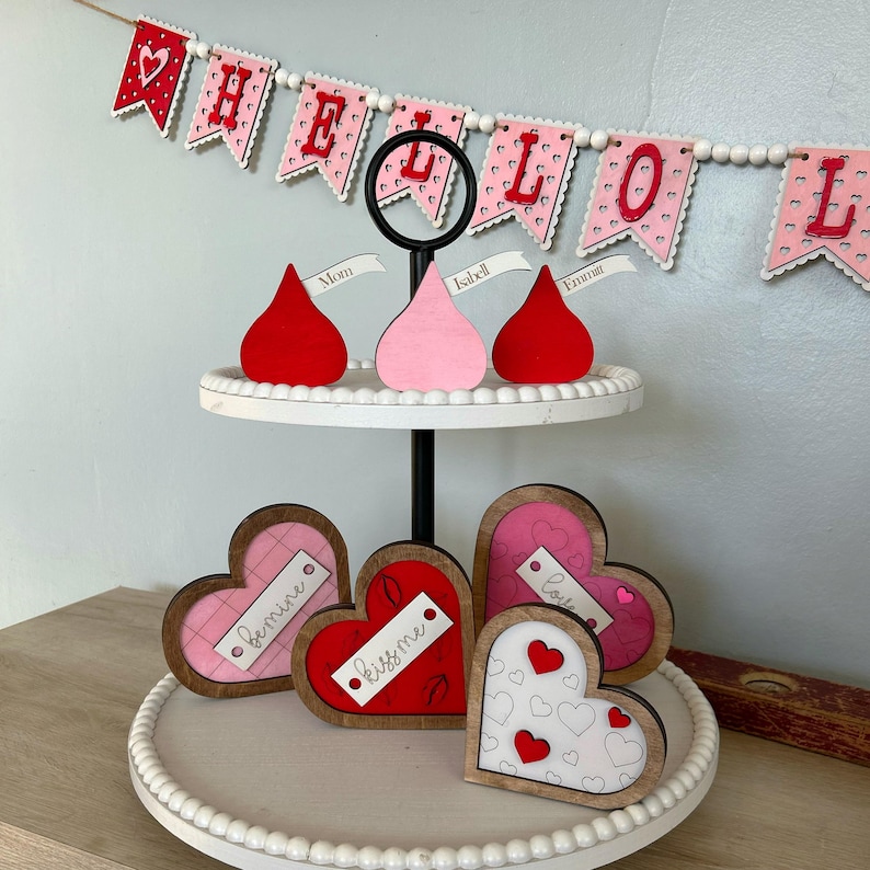 May include: A two-tiered white tiered tray with a black metal stand. The top tier has three red, pink, and white wooden heart-shaped ornaments with the words "Mom", "Isabel", and "Emma" written on them. The bottom tier has four wooden heart-shaped ornaments with the words "Love", "Kiss Me", "Bemine", and a heart with a pattern of small hearts. The tray is decorated with white pearl beads.