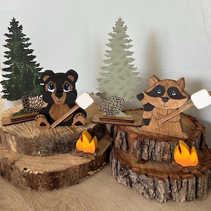 May include: Wooden figurines of a bear and raccoon roasting marshmallows over a campfire. The figurines are painted in brown, black, and gray. The campfire is represented by orange flames on a wooden base.