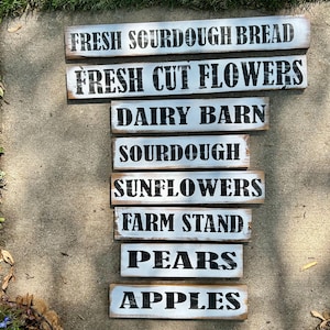 Puede incluir: Una colecci&oacute;n de letreros r&uacute;sticos de madera con letras negras sobre un fondo blanco y azul desgastado. Los letreros dicen "FRESH SOURDOUGH BREAD", "FRESH CUT FLOWERS", "DAIRY BARN", "SOURDOUGH", "SUNFLOWERS", "FARM STAND", "PEARS" y "APPLES".