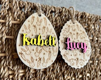 May include: Two wooden Easter egg ornaments with bunny and carrot designs. The top ornament is oval shaped and has the name "Isabelle" written in yellow letters. The bottom ornament is round and has the name "Lucy" written in pink letters.