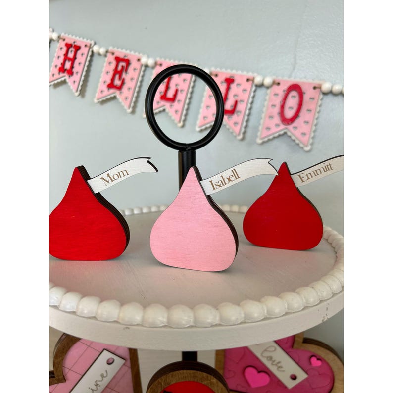 May include: A tiered tray decorated with red and pink heart-shaped decorations and a banner that says "HELLO". There are three wooden chocolate kiss-shaped place card holders with the names "Mom", "Isabell", and "Emmitt" written on them.