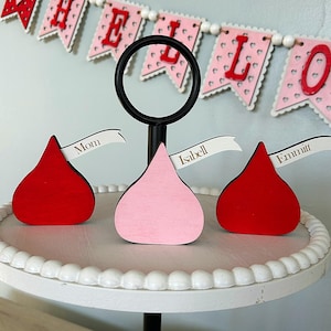 May include: Three red and pink wooden kiss-shaped place card holders with white flags that say "Mom", "Isabel", and "Emmitt". The place card holders are on a white tiered cake stand.