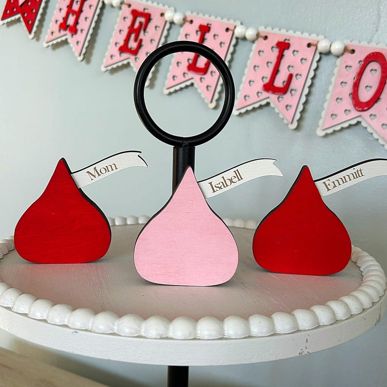 May include: Three red and pink wooden kiss-shaped place card holders with white flags that say "Mom", "Isabel", and "Emmitt". The place card holders are on a white tiered cake stand.