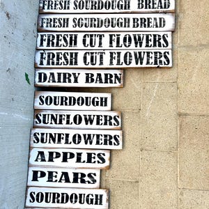 Puede incluir: Una colecci&oacute;n de letreros r&uacute;sticos de madera pintados de blanco con letras negras. Los letreros anuncian productos frescos de la granja, incluyendo "Pan de masa madre fresco", "Flores frescas cortadas", "Granja lechera", "Girasoles", "Manzanas" y "Peras".