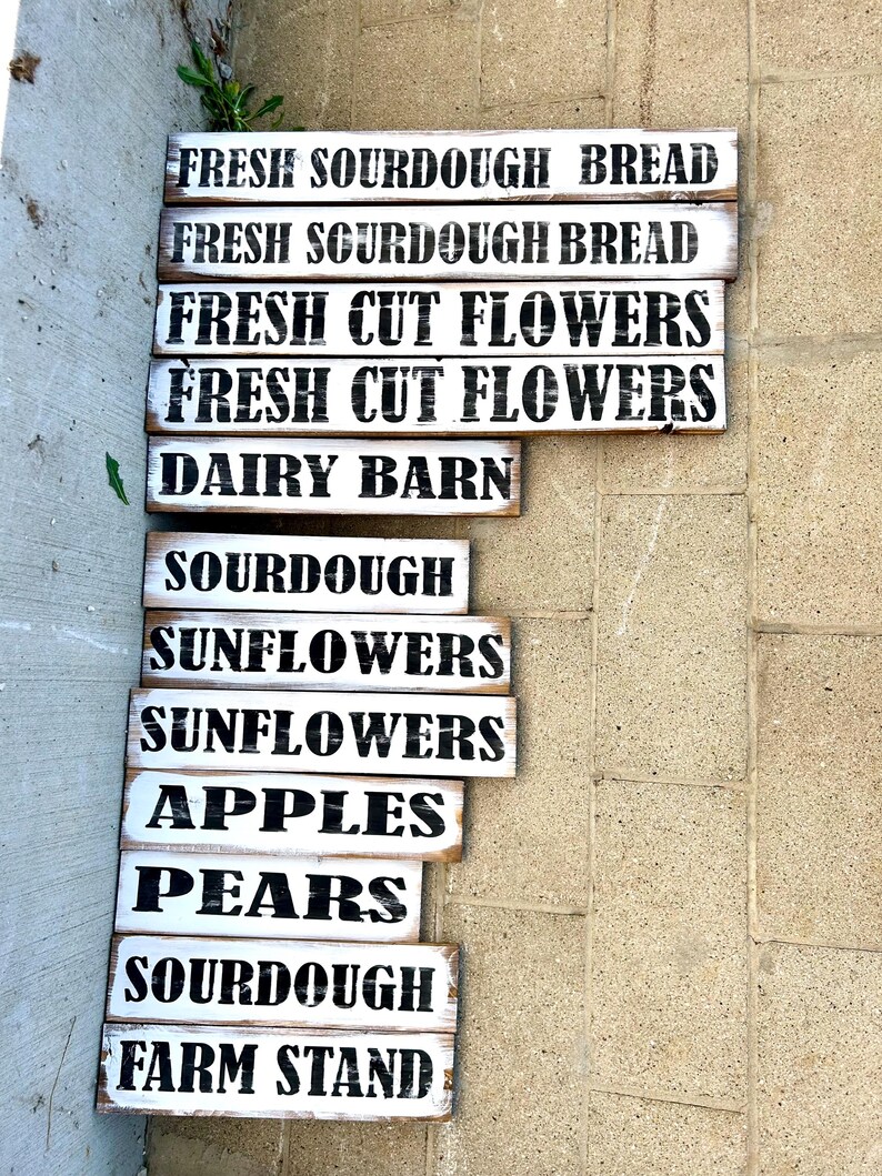 Puede incluir: Una colecci&oacute;n de letreros r&uacute;sticos de madera pintados de blanco con letras negras. Los letreros anuncian productos frescos de la granja, incluyendo "Pan de masa madre fresco", "Flores frescas cortadas", "Granja lechera", "Girasoles", "Manzanas" y "Peras".