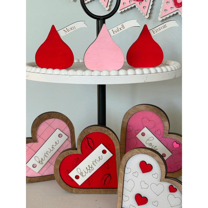 May include: Three wooden heart-shaped decorations with different designs and sayings. The first heart is pink with a white background and the words "be mine" written on it. The second heart is red with a white background and the words "kiss me" written on it. The third heart is white with a black background and a pattern of small hearts. The hearts are on a tiered stand with three wooden chocolate kiss decorations on the top tier. The chocolate kisses are red, pink, and red and have names written on them.