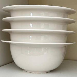 Op de afbeelding: Een stapel van vier witte keramische kommen. Elke kom heeft een afgeronde basis en een brede, platte rand. De kommen zijn netjes op elkaar gestapeld, waarbij de randen van elke kom een gelaagd effect cre&euml;ren. De kommen hebben een glanzende afwerking.