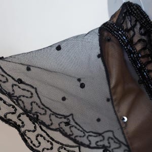 May include: Close-up of a black sheer garment with a delicate, dotted pattern. The edges are embellished with intricate beadwork and a decorative, swirling design. The garment appears to be layered over a tan fabric.