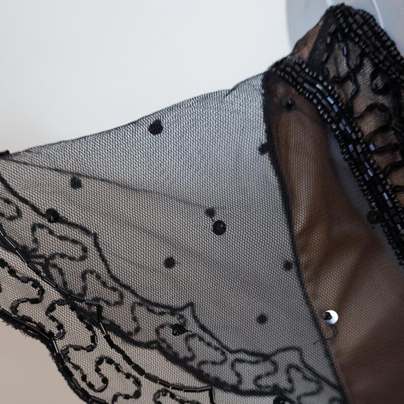 May include: Close-up of a black sheer garment with a delicate, dotted pattern. The edges are embellished with intricate beadwork and a decorative, swirling design. The garment appears to be layered over a tan fabric.
