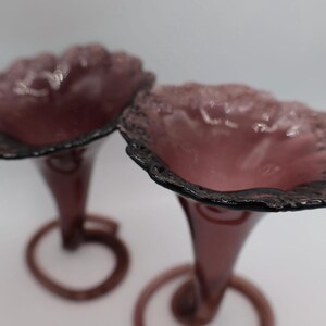 Könnte beinhalten: Zwei antike Vasen aus amethystfarbenem Glas. Jede Vase hat einen ausgestellten, gerüschten Rand mit dunklem, strukturiertem Rand. Die Stiele sind schlank und münden in einen kreisförmigen Sockel. Die Vasen sind wahrscheinlich dekorativ.