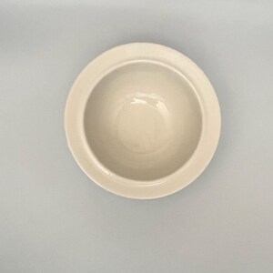 Op de afbeelding: Een cr&egrave;mekleurige keramische kom met een brede rand. De kom heeft een eenvoudig, klassiek ontwerp en lijkt leeg te zijn. De kom is gecentreerd in het frame tegen een lichtgrijze achtergrond.