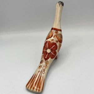 May include: A hand-painted, decorative bird figurine. The bird has a cream-colored body with a long neck and tail. It features a large red flower design on its body, with brown and orange accents. The bird stands on three small legs.