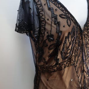 May include: A black beaded dress with a V-neckline and short sleeves. The dress features intricate sequin and bead detailing on a sheer overlay, with a tan underlay. The design includes floral and geometric patterns.