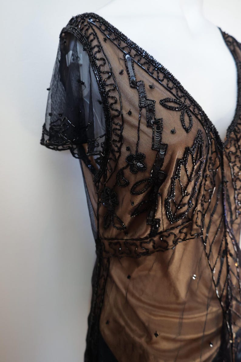 May include: A black beaded dress with a V-neckline and short sleeves. The dress features intricate sequin and bead detailing on a sheer overlay, with a tan underlay. The design includes floral and geometric patterns.