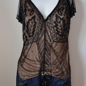 May include: A black beaded top with a sheer overlay and a V-neckline. The top features intricate leaf and geometric patterns, with a tan underlay visible beneath the sheer fabric. The short sleeves and hemline are also trimmed with black beading.