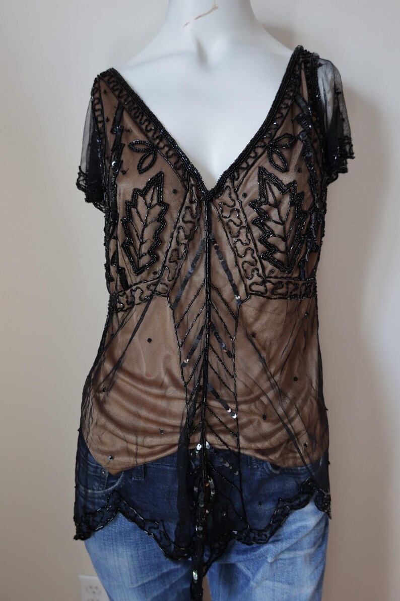 May include: A black beaded top with a sheer overlay and a V-neckline. The top features intricate leaf and geometric patterns, with a tan underlay visible beneath the sheer fabric. The short sleeves and hemline are also trimmed with black beading.