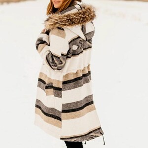 May include: A long, striped coat with a hood and faux fur trim. The coat features horizontal stripes in cream, black, and tan. The hood is lined with faux fur. The coat has a drawstring waist and is worn in a snowy environment.