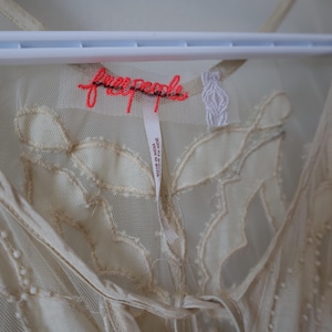 Puede incluir: Vestido transparente color crema con patrones bordados en forma de hojas. El vestido est&aacute; colgado de una percha blanca. La marca "free people" est&aacute; bordada en color coral. Una etiqueta dice "Made in India".