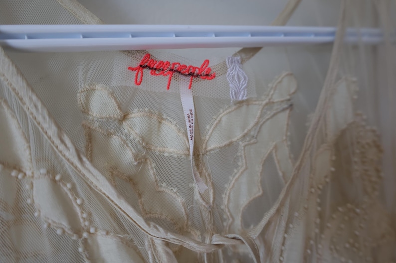 Puede incluir: Vestido transparente color crema con patrones bordados en forma de hojas. El vestido est&aacute; colgado de una percha blanca. La marca "free people" est&aacute; bordada en color coral. Una etiqueta dice "Made in India".