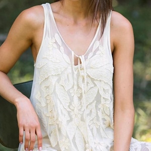 Puede incluir: Vestido sin mangas de color marfil con intrincados detalles de encaje floral. El vestido presenta un escote en V con un cierre de lazo y una delicada borla. La tela es transparente, creando un efecto de capas.