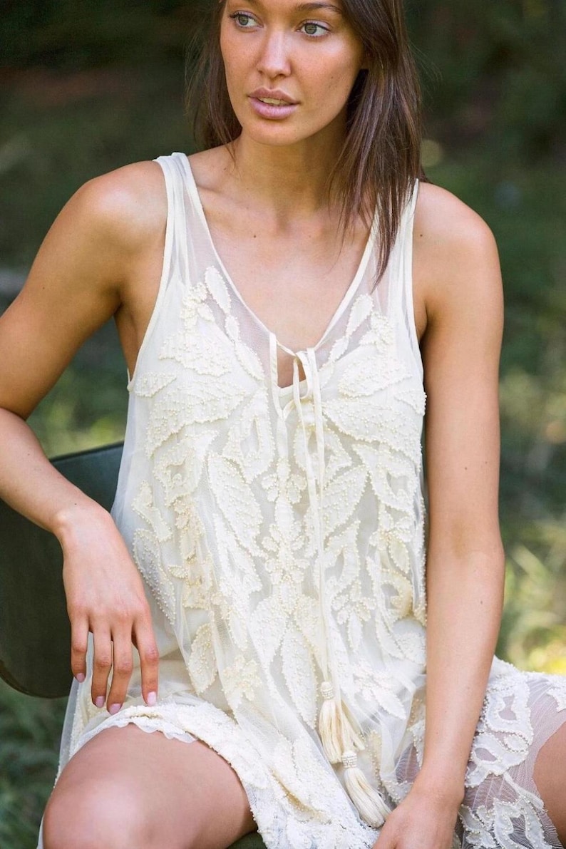 Puede incluir: Vestido sin mangas de color marfil con intrincados detalles de encaje floral. El vestido presenta un escote en V con un cierre de lazo y una delicada borla. La tela es transparente, creando un efecto de capas.