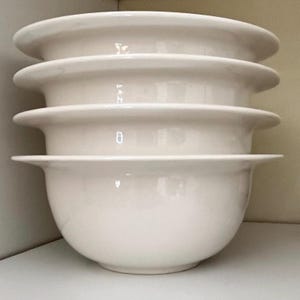 Op de afbeelding: Een stapel van vier witte keramische kommen. Elke kom heeft een afgeronde basis en een brede, platte rand. De kommen zijn op elkaar gestapeld, met de randen naar buiten gericht. De kommen zijn effen, gebroken wit van kleur.