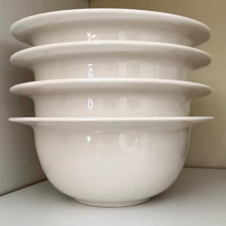 Op de afbeelding: Een stapel van vier witte keramische kommen. Elke kom heeft een afgeronde basis en een brede, platte rand. De kommen zijn op elkaar gestapeld, met de randen naar buiten gericht. De kommen zijn effen, gebroken wit van kleur.