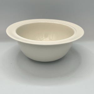 Op de afbeelding: Een cr&egrave;mekleurige keramische kom met een brede, platte rand. De kom heeft een afgeronde vorm met een gladde, glanzende afwerking. De binnenkant van de kom is iets verzonken. De kom is ontworpen voor het serveren van voedsel.