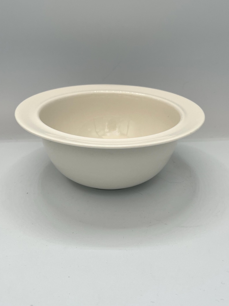 Op de afbeelding: Een cr&egrave;mekleurige keramische kom met een brede, platte rand. De kom heeft een afgeronde vorm met een gladde, glanzende afwerking. De binnenkant van de kom is iets verzonken. De kom is ontworpen voor het serveren van voedsel.