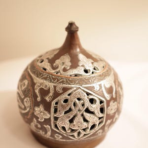 May include: A brown metal decorative container with intricate silver filigree patterns. The container has a rounded shape with a small, pointed top and a base. The design includes floral and geometric elements.