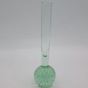 May include: A clear glass vase with a long, slender neck and a round, green base. The base is textured with small bubbles. The vase is designed for a single stem flower or a small floral arrangement.