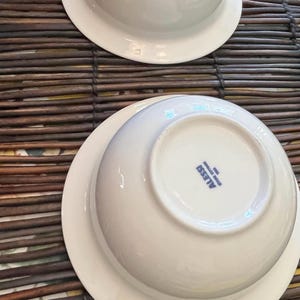 Op de afbeelding: Twee witte keramische kommen met een brede rand, op een bruine bamboemat. De kommen zijn ondersteboven en tonen de merknaam "ALESSI" in blauwe letters op de bodem. De kommen hebben een strak design.