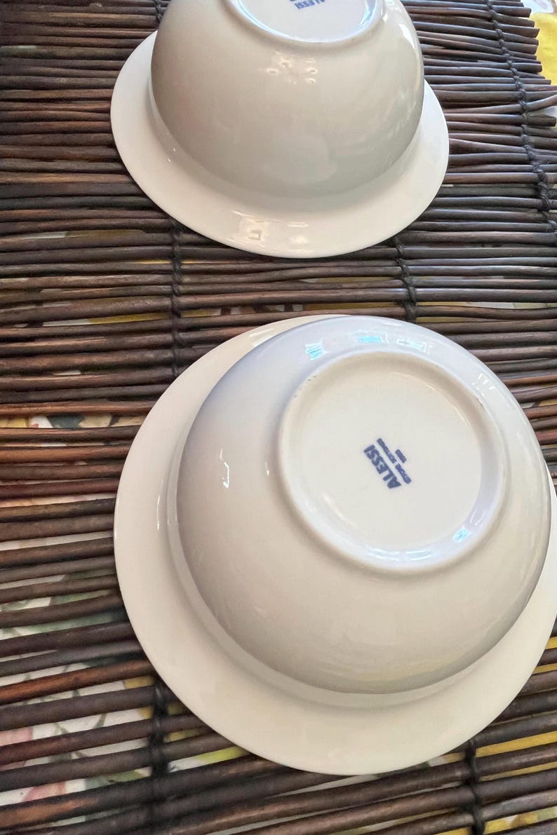 Op de afbeelding: Twee witte keramische kommen met een brede rand, op een bruine bamboemat. De kommen zijn ondersteboven en tonen de merknaam "ALESSI" in blauwe letters op de bodem. De kommen hebben een strak design.