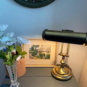May include: A brass desk lamp with a black shade and a framed watercolor painting of a cottage in a green landscape. The painting is on a dark gray table with a vase of white flowers.