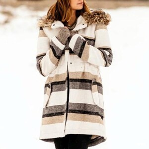 May include: A winter coat with a striped pattern in shades of white, brown, and black. The coat features a fur-lined hood and pockets. The person is wearing matching gloves. The coat is designed for cold weather.