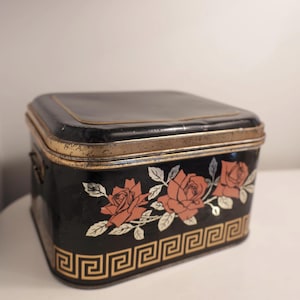 Può includere: Una scatola vintage in metallo nero con coperchio incernierato. La scatola è decorata con rose arancioni e dettagli dorati. Un motivo a chiave greca dorata borda il bordo inferiore. La scatola ha una forma rettangolare.