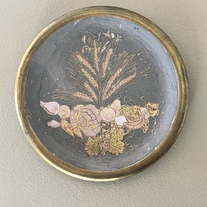 Puede incluir: Plato decorativo redondo con fondo gris oscuro y borde dorado. El plato presenta un diseño floral y de trigo en tonos rosa, dorado y verde. El plato es probablemente de metal.
