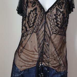 May include: Black beaded top with a V-neckline and short sleeves. The sheer overlay features leaf and geometric patterns. The top is layered over a nude-colored fabric and has a scalloped hemline.