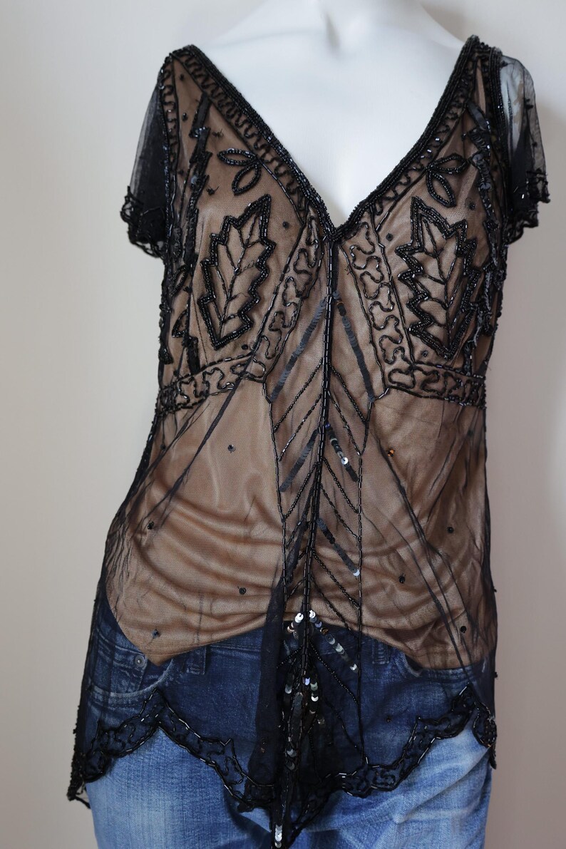 May include: Black beaded top with a V-neckline and short sleeves. The sheer overlay features leaf and geometric patterns. The top is layered over a nude-colored fabric and has a scalloped hemline.