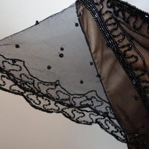 May include: Black beaded evening wear with a sheer overlay. The garment features intricate beadwork along the neckline and sleeve edges, with a delicate, dotted mesh fabric. The design includes a scalloped hem.