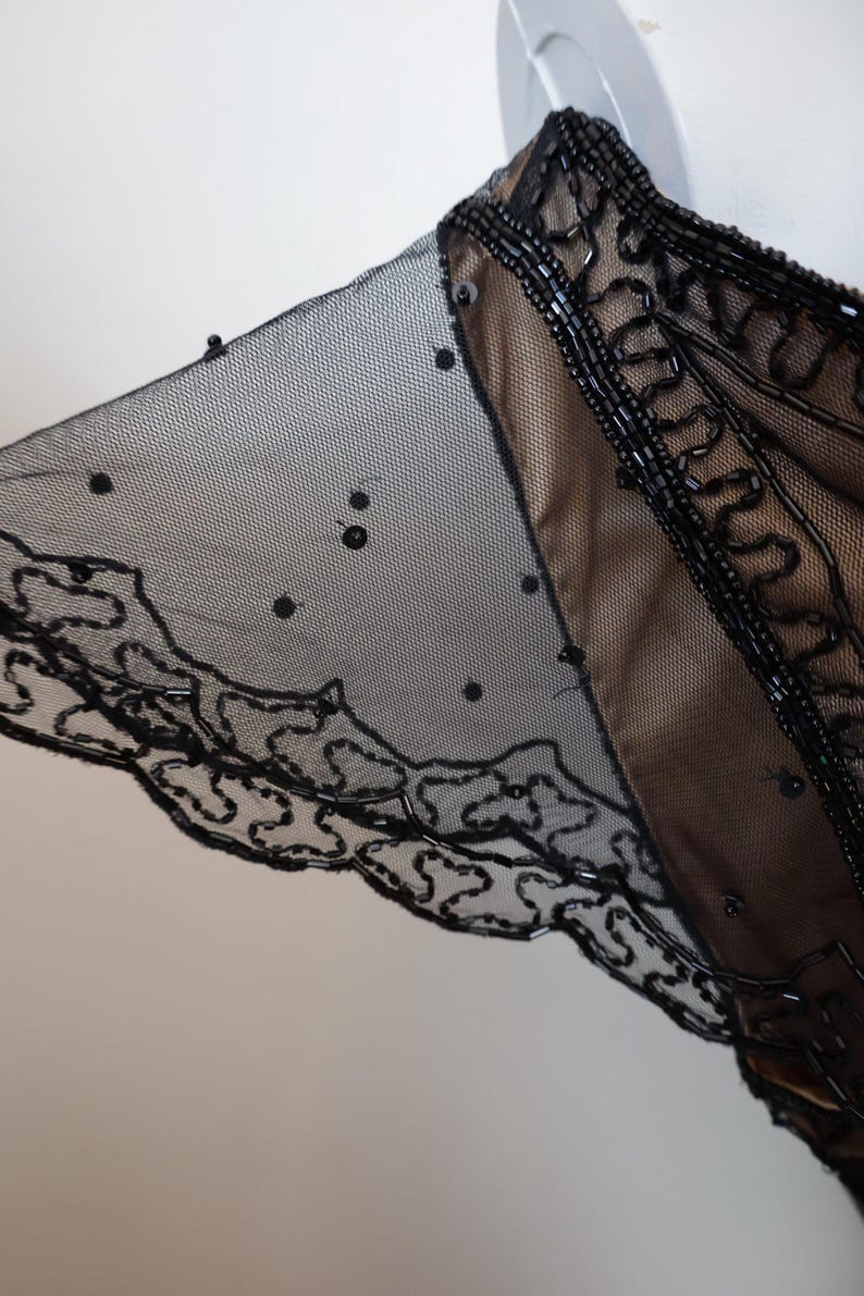 May include: Black beaded evening wear with a sheer overlay. The garment features intricate beadwork along the neckline and sleeve edges, with a delicate, dotted mesh fabric. The design includes a scalloped hem.