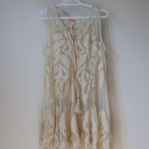 Puede incluir: Vestido sin mangas color crema con intrincada bordado de hojas. El vestido presenta un escote en V, un lazo con borla y una capa transparente. Cuelga de una percha blanca contra una pared blanca lisa, mostrando su delicado dise&ntilde;o.