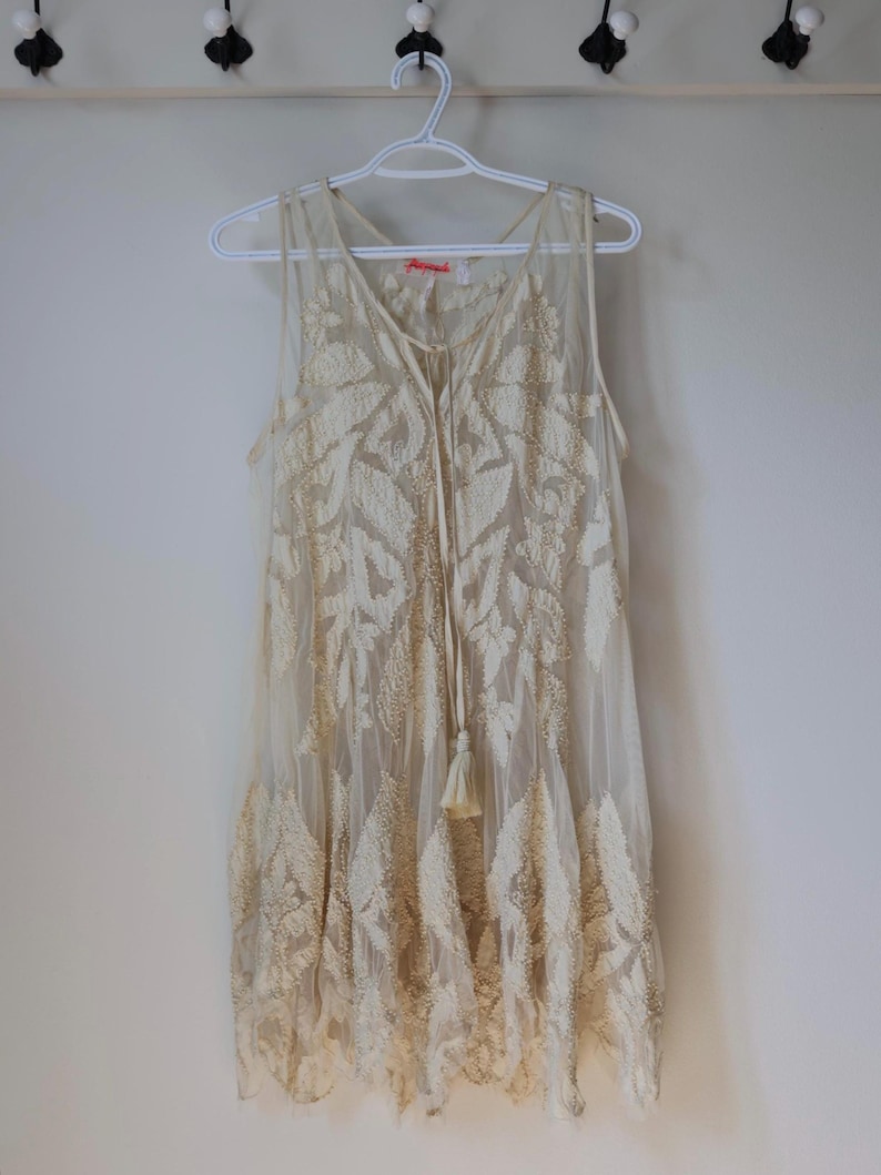 Puede incluir: Vestido sin mangas color crema con intrincada bordado de hojas. El vestido presenta un escote en V, un lazo con borla y una capa transparente. Cuelga de una percha blanca contra una pared blanca lisa, mostrando su delicado dise&ntilde;o.