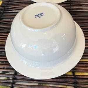 Op de afbeelding: Twee witte porseleinen kommen met bijpassende schotels. De kommen staan ondersteboven en onthullen de merknaam "Alessi" in blauwe letters. De kommen en schotels staan op een bruin en geel geweven oppervlak.