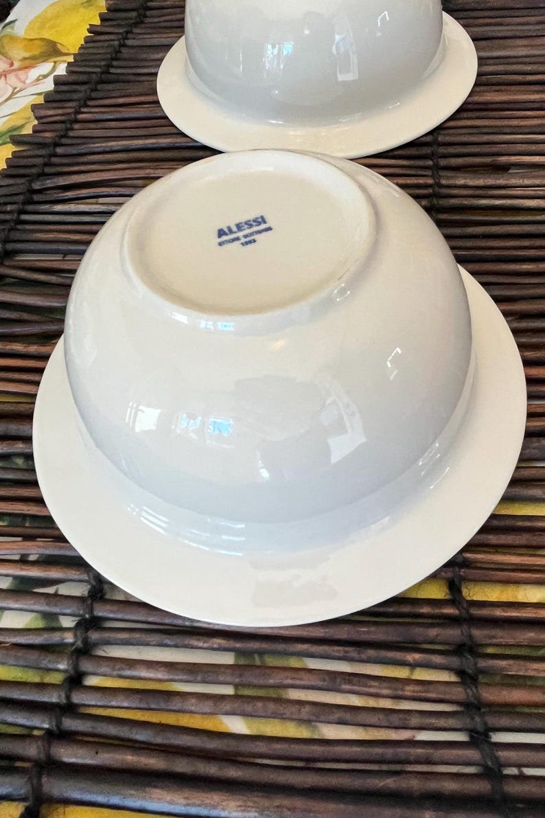 Op de afbeelding: Twee witte porseleinen kommen met bijpassende schotels. De kommen staan ondersteboven en onthullen de merknaam "Alessi" in blauwe letters. De kommen en schotels staan op een bruin en geel geweven oppervlak.