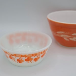 May include: Two vintage mixing bowls. One is white with an orange floral pattern around the rim. The other is orange with a white wheat design. Both bowls are made of glass and are set against a white background.