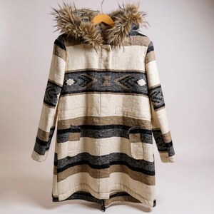 May include: A long, striped coat with a faux fur-lined hood. The coat features a pattern of black, gray, and white geometric shapes. It has two front pockets and a zipper closure. The coat is hanging on a wooden hanger.