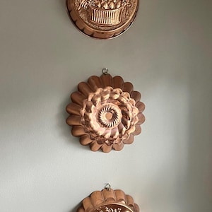 May include: Three circular copper molds arranged vertically. The top and bottom molds feature embossed fruit basket designs, while the center mold has a fluted, scalloped edge with a central spiral pattern. Each mold includes a small metal hanging loop.